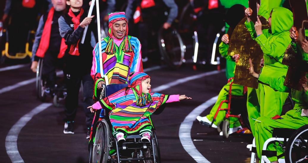 Un hombre lleva a una niña en silla de ruedas, ambos sonrientes, frente a la delegación Parapanamericana peruana en la Ceremonia de Inauguración de los Juegos Parapanamericanos Lima 2019 en el Estadio Nacional
