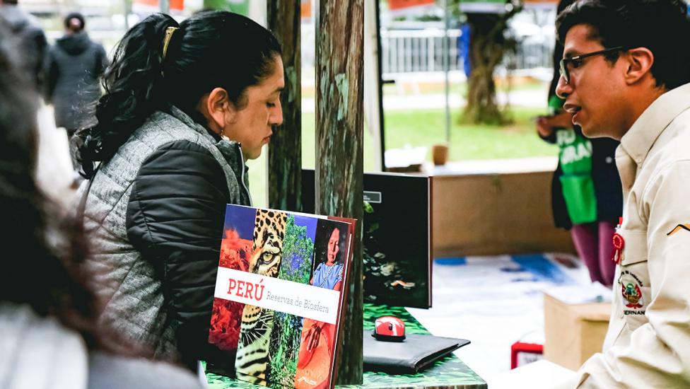 Voluntario de Sernanp informa a mujer que se acerca al módulo donde se encuentra la calculadora de la huella de carbono, en Lima 2019.